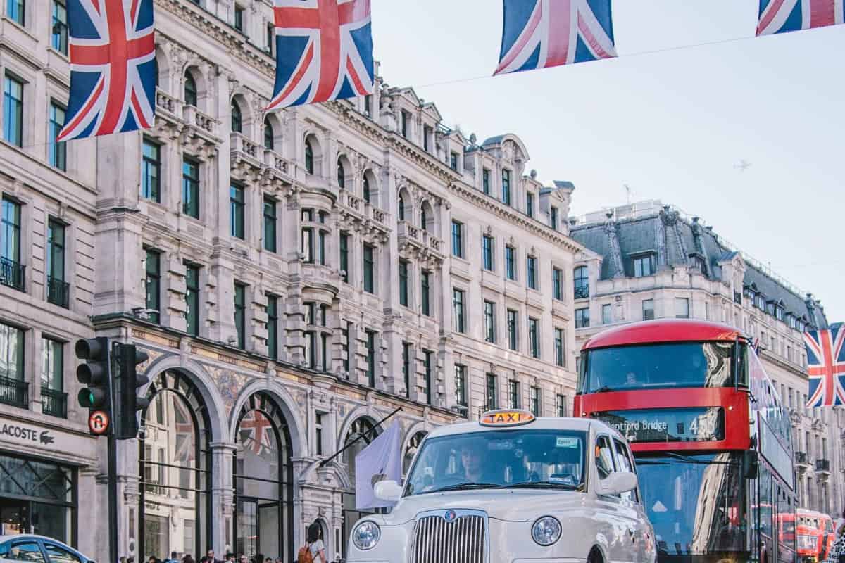 A lively London street features a classic white taxi and a red double-decker bus. Union Jack flags flutter overhead, and historic buildings line the road beneath a clear blue sky, capturing the dynamic spirit often associated with the bustling Stock Market scene.