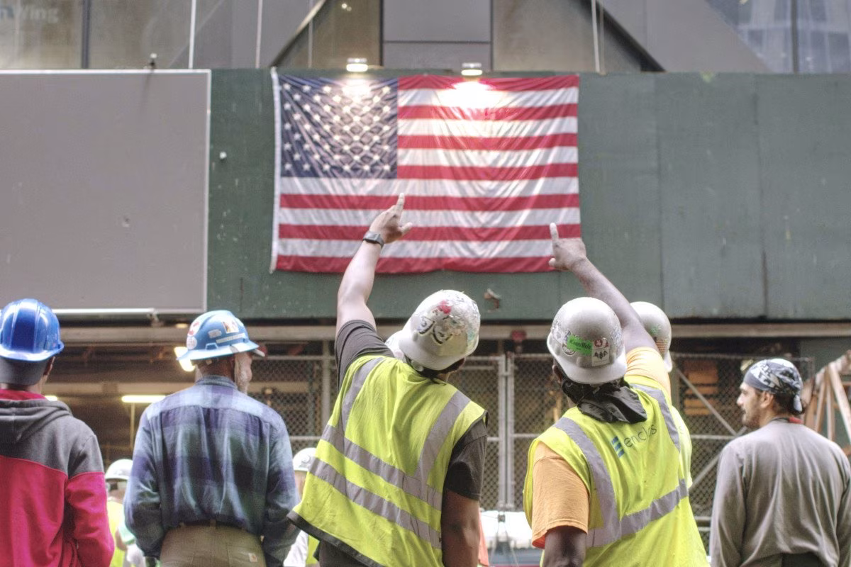 Construction workers in hard hats look at an American flag on a building site. from Stockmark.it