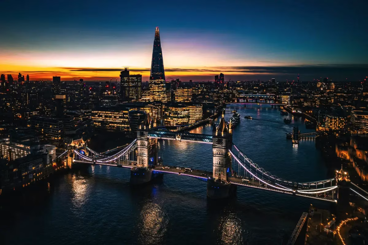 Aerial view of London’s Tower Bridge illuminated at dusk, with city lights reflecting on the River Thames and The Shard skyscraper—home to innovative offices like Amazon and Google—rising in the background under a vibrant sunset sky. @ stockmark.it