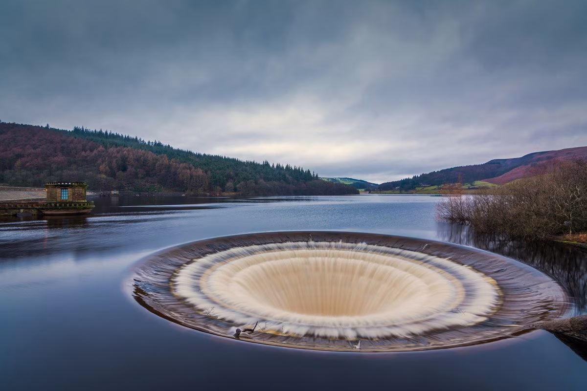 A large circular overflow drain in a calm reservoir, operated by Severn Trent, is surrounded by trees and hills under a cloudy sky. @ stockmark.it