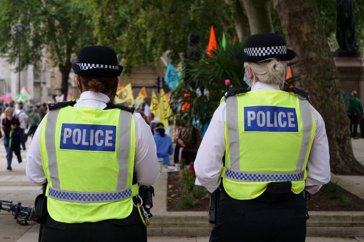 Two police officers in high-visibility jackets stand with their backs to the camera at a public event with people, flags, and trees in the background, highlighting the presence of Met police jobs at such gatherings. @ stockmark.it