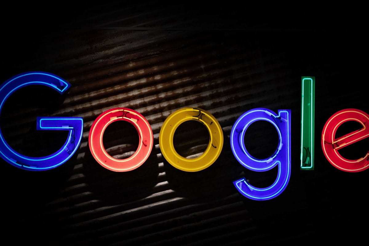 A brightly illuminated Google logo in neon colors—blue, red, yellow, green, and light blue—shines against a dark, textured background. The vibrant display evokes the dynamic and ever-changing nature of the stock market.